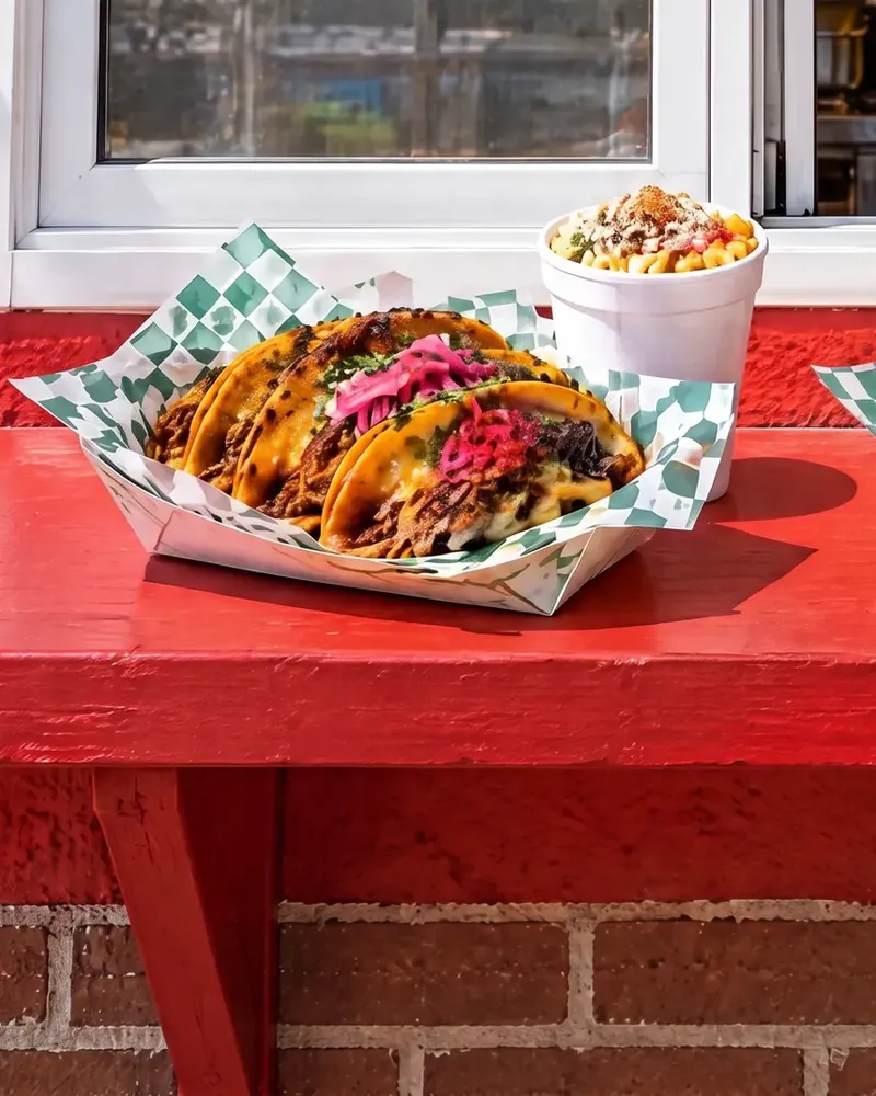 Queso birria tacos in a paper-lined tray beside a cup of esquite, on a red counter.